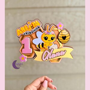 Puede incluir: Adorno para tarta con una abeja de dibujos animados, el texto "Abejita Chiquitita" y el nombre "Alana". El dise&ntilde;o incluye un n&uacute;mero 1, flores y un fondo de panal. El adorno es en tonos rosa, amarillo y dorado.