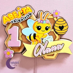 Puede incluir: Adorno de pastel en capas con una abeja de dibujos animados con una corona de flores, un dise&ntilde;o de panal y el nombre "Alana". El adorno incluye el texto "Abeja Chiquitita" y el n&uacute;mero "1", con detalles en rosa y amarillo.