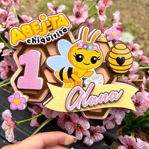 Puede incluir: Un adorno para pastel colorido con una abeja de dibujos animados, el texto "Abeijita Chiquitita" y el nombre "Alana". El adorno incluye el n&uacute;mero "1" y un dise&ntilde;o de panal, con detalles en rosa y amarillo. El fondo est&aacute; borroso con flores rosas.