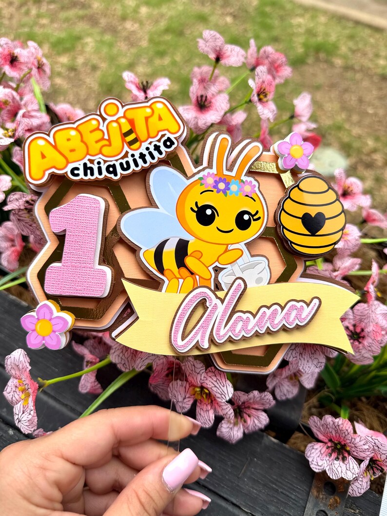 Puede incluir: Un adorno para pastel colorido con una abeja de dibujos animados, el texto "Abeijita Chiquitita" y el nombre "Alana". El adorno incluye el n&uacute;mero "1" y un dise&ntilde;o de panal, con detalles en rosa y amarillo. El fondo est&aacute; borroso con flores rosas.