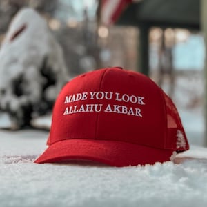 May include: Red baseball cap with a white mesh back. The cap has white embroidered text that reads "Made You Look Allahu Akbar".