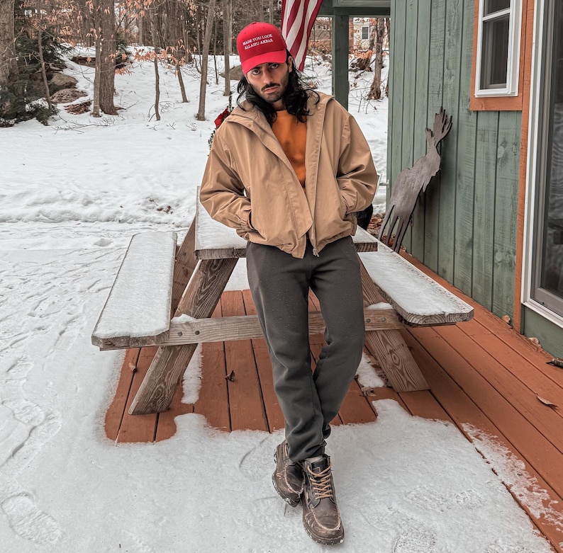 May include: A person wearing a red baseball cap with the text "Make America Great Again" in white letters, a tan jacket, and dark grey pants. The person is standing on a wooden deck with snow on the ground.