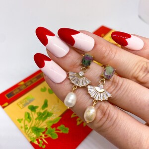May include: Gold earrings with a green stone and a pearl drop. The earrings feature a fan-shaped design with clear rhinestones.
