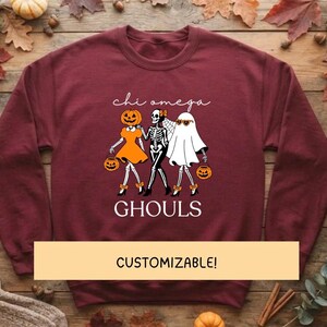 Könnte beinhalten: Ein bordeauxrotes Langarm-Sweatshirt mit einer Grafik von drei Figuren: eine Kürbiskopf-Figur in einem orangefarbenen Kleid, ein Skelett und ein Geist mit Sonnenbrille. Der Text lautet "chi omega" und "GHOULS". Ein gelbes Banner zeigt "CUSTOMIZABLE!"