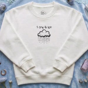 May include: A white long-sleeve crewneck sweatshirt featuring a black graphic of a rain cloud with the text "I cry a lot" printed above it. The garment has ribbed cuffs and a ribbed hem, with a small circular logo printed on the inner neck area.