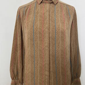 May include: A long-sleeved button-up shirt in a tan color featuring a dark brown herringbone pattern. The shirt is accented with vertical pinstripes in alternating shades of light blue and pink, and it includes a standard collar and buttoned cuffs.