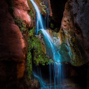 May include: A cascading waterfall flows over moss-covered rocks in a dark, narrow canyon. The water is a vibrant blue, and the rocks are a rich red and brown.