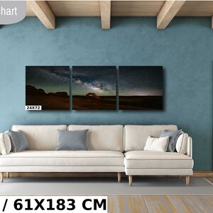 May include: A three-piece canvas print set featuring a night sky with stars and the Milky Way over a desert landscape with a natural archway. The print set is displayed on a wall above a white sectional sofa in a living room setting. The text "24X72 IN / 61X183 CM" is displayed below the image.