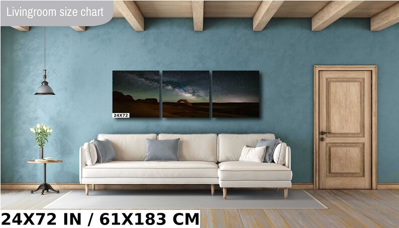 May include: A three-piece canvas print set featuring a night sky with stars and the Milky Way over a desert landscape with a natural archway. The print set is displayed on a wall above a white sectional sofa in a living room setting. The text "24X72 IN / 61X183 CM" is displayed below the image.