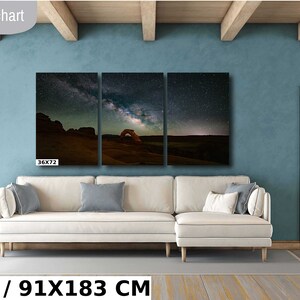 May include: A three-panel canvas print of a starry night sky with the Milky Way galaxy over a red rock arch. The print is 36 inches by 72 inches and is displayed on a white sectional sofa in a living room with a blue wall.