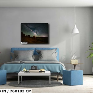 May include: A bedroom interior with a blue bed, two blue ottomans, and a coffee table. A framed photograph of a starry night sky with a red rock archway hangs on the wall above the bed. The photograph is 30x40 inches.