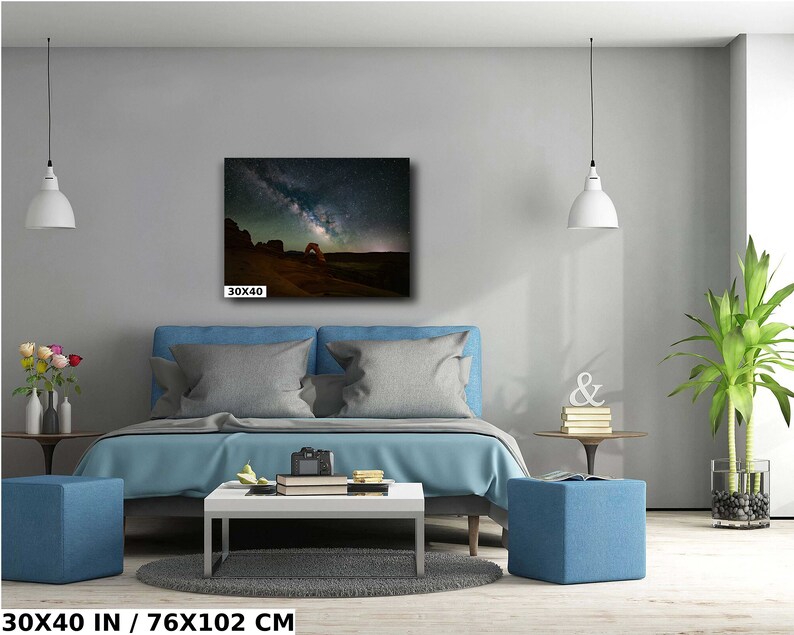 May include: A bedroom interior with a blue bed, two blue ottomans, and a coffee table. A framed photograph of a starry night sky with a red rock archway hangs on the wall above the bed. The photograph is 30x40 inches.
