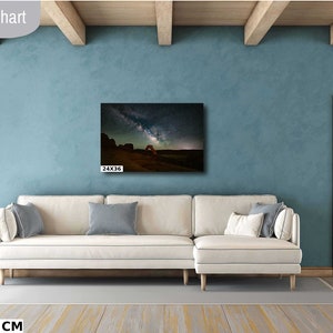 May include: A white sectional sofa with two pillows in a living room with a blue wall and a wooden door. A canvas print of a starry night sky with a rock archway is hanging on the wall above the sofa. The print is 24x36 inches.