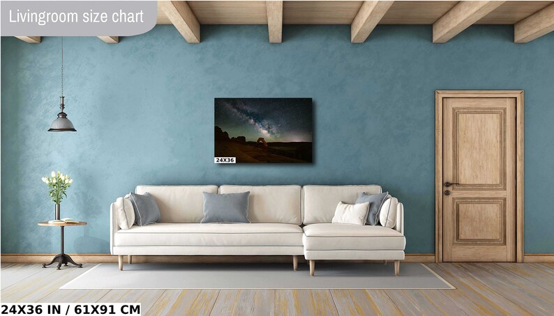 May include: A white sectional sofa with two pillows in a living room with a blue wall and a wooden door. A canvas print of a starry night sky with a rock archway is hanging on the wall above the sofa. The print is 24x36 inches.