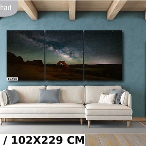 May include: A three-panel canvas print of a night sky with the Milky Way galaxy over a desert landscape. The print is 40 inches by 90 inches and is displayed on a wall in a living room setting. The text "40X90 IN / 102X229 CM" is displayed below the print.