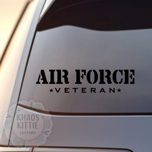 May include: Black vinyl decal with the words "AIR FORCE" above "*VETERAN*" in a bold, stencil-style font. The decal is affixed to a car window. The decal also includes the text "KHAOS KITTIE CUSTOMS".