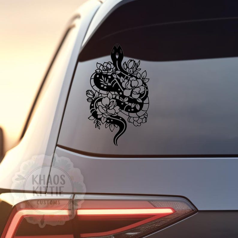 May include: Black vinyl car decal featuring a snake intertwined with floral elements. The snake's body is detailed with dots, and the flowers have intricate petal designs. The decal is applied to a car window, with the text "KHAOS KITTIE CUSTOMS" visible.
