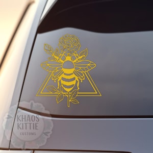 May include: A yellow vinyl decal applied to a car window, featuring a line art illustration of a bee centred within a triangle, topped with a rose and surrounded by leaves. A watermark in the corner displays the text KHAOS KITTIE CUSTOMS.