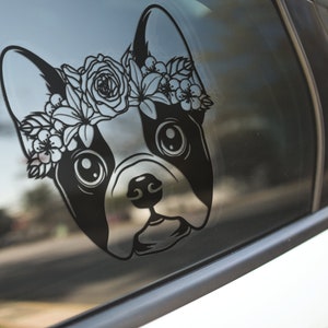 May include: Black and white silhouette of a French bulldog wearing a floral crown. The dog is looking to the right. The image is on a car window.