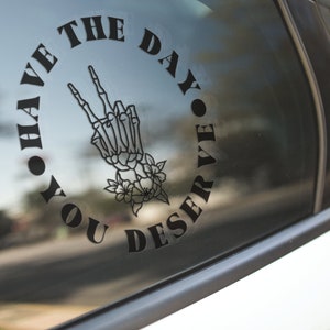 May include: Black decal on a car window featuring a skeleton hand making a peace sign, surrounded by the words "YOU HAVE THE DAY YOU DESERVE" in a circular arrangement. The hand is adorned with floral accents.