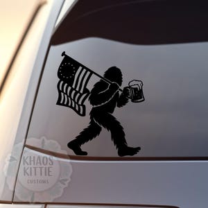 May include: Black vinyl decal of a Bigfoot carrying an American flag and a beer stein. The Bigfoot is walking to the right. The decal is applied to a car window. The text "Khaos Kittie Customs" is in the lower left corner.