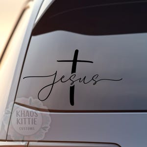 May include: Black decal of a cross with the word "Jesus" in cursive script. The decal is affixed to a car window. A logo with the text "KHAOS KITTIE CUSTOMS" is visible in the lower left corner.