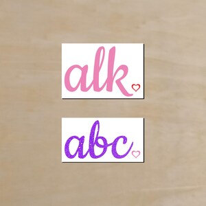 May include: Two rectangular white cards with cursive text and heart accents. The top card reads "alk" in pink, and the bottom card reads "abc" in purple glitter. Both cards have a small heart.