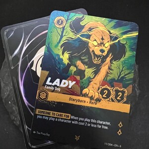 May include: Two trading cards. The front card features a snarling dog with glowing yellow eyes, titled "LADY Family Dog." The card has a gold and black border with the text "Storyborn Hero." The other card is black with white and purple designs.