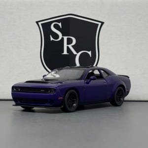 May include: A dark blue diecast model car with black wheels. The car is a coupe with a sleek design. The background features a white and black logo. The car is a miniature replica of a sports car.