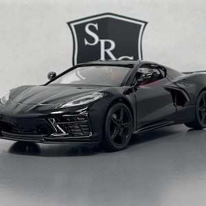 May include: A black die-cast model sports car featuring a sleek aerodynamic body, sharp angular headlights, five-spoke black wheels, and side air intakes. The vehicle is positioned in front of a white background with a black shield logo containing the letters SR.