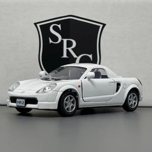 May include: White die-cast model car of a Toyota MR2, a two-seater sports car with a black interior and black wheels. The car has a black license plate that reads "MR-2".