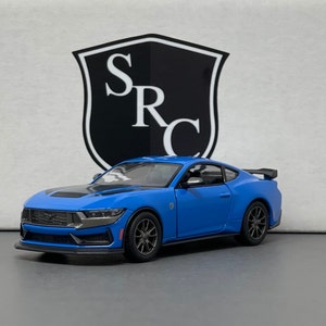 May include: A blue diecast model car of a Ford Mustang with a black spoiler and black accents. The car is sitting on a white surface.