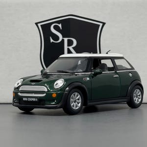 Op de afbeelding: Een donkergroene Mini Cooper S modelauto met een wit dak. De auto heeft zwarte banden en zilveren velgen. De woorden "MINI COOPER S" staan op de voorkant. Een zwart-wit logo staat op de achtergrond.