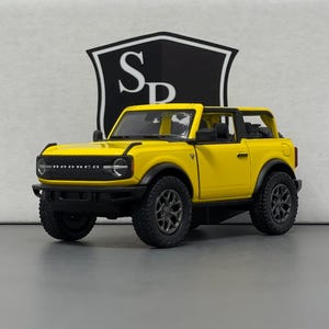 May include: A yellow toy SUV with black wheels and trim. The vehicle has a black front grill and a black roll bar. The background features a black and white logo.