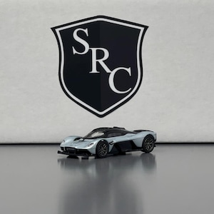 May include: A silver and black toy car model of a McLaren Speedtail. The car is on a white surface with a black shield logo that says "SRC" in white letters.