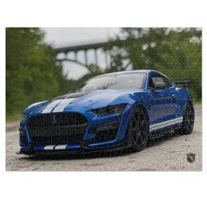 Może przedstawiać: Niebieski Ford Mustang Shelby GT500 z białymi paskami wyścigowymi zaparkowany na szarej powierzchni asfaltowej. Samochód jest skierowany przodem do kamery, widoczne są przedni grill i reflektory. Tło jest rozmyte i przedstawia zielone drzewa oraz most.