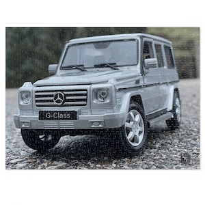 Könnte beinhalten: Puzzle eines silbernen G-Klasse SUV. Das Puzzle zeigt eine detaillierte Nachbildung des Fahrzeugs, einschließlich des Mercedes-Benz-Logos und des Textes "G-Klasse" auf der Vorderseite. Das Puzzle liegt auf einer Schotteroberfläche.