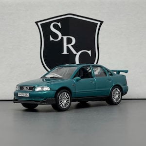 May include: A teal Audi A4 model car with silver rims and a rear spoiler. The car has a license plate that reads "HHNK282" and the Audi logo on the front. The background features a white and black shield logo with the letters "SRC".