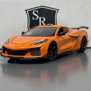 Può includere: Modello di auto sportiva arancione con ruote nere e spoiler posteriore. L'auto ha un design elegante con fari dettagliati e una griglia anteriore nera. Il modello è ambientato su uno sfondo neutro con un logo.