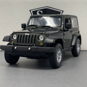 Könnte beinhalten: Ein dunkelgrünes Jeep Rubicon Modell mit schwarzen Akzenten. Das Modell zeigt das Jeep-Logo vorne und Rubicon an der Seite. Die Reifen sind schwarz und die Felgen silbern. Es ist eine Miniatur-Nachbildung des ikonischen Geländewagens.