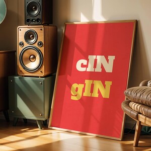 Op de afbeelding: Een kamer in retrostijl met een rood omkaderde poster met de woorden "cIN gIN" in wit en geel. Een bruine leren fauteuil met een deken staat naast de poster. Luidsprekers en een vintage radio staan op een houten kast.