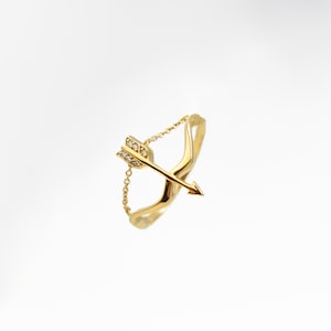 May include: A gold ring with a delicate chain and a bow and arrow design with small clear stones.