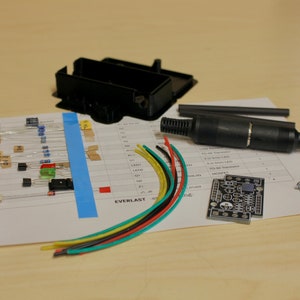 Pode incluir: Um kit para um projeto de eletrónica, contendo uma caixa de plástico preto, uma placa de circuito impresso, uma variedade de componentes eletrónicos, incluindo resistências, capacitores e LEDs, um conector preto, um conjunto de parafusos e cinco fios de diferentes cores: vermelho, preto, amarelo, verde e azul.