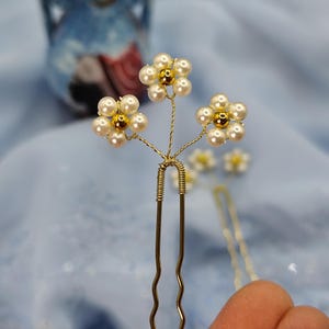 May include: Gold hair pins with three white pearl flowers with gold centers. The flowers are arranged in a cluster on the top of the pins.