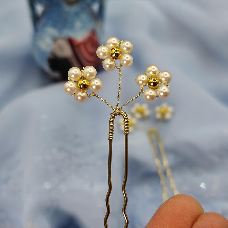 May include: Gold hair pins with three white pearl flowers with gold centers. The flowers are arranged in a cluster on the top of the pins.