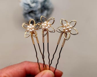 May include: Three gold-coloured hair pins with flower-shaped designs. Each flower has five petals and a cluster of small, white pearls at the centre. The pins are held in a hand, with a blurred blue object in the background. These hair accessories are ideal for weddings or special occasions.