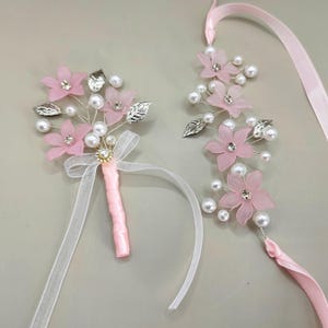 May include: A set of two pink flower boutonnieres with pearl accents and silver leaves. The boutonnieres are attached to pink ribbons.