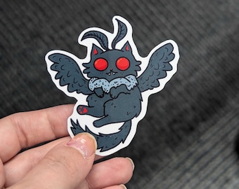May include: A black and gray sticker of a cat with wings and red eyes. The cat is wearing a white fluffy collar.