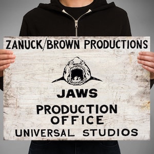 May include: A white wooden sign with black text that reads "Zanuck/Brown Productions", a black and white image of a shark, "Jaws", "Production Office", and "Universal Studios".