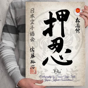 May include: A black and white calligraphy print of the Japanese word "Oss" meaning "Yes Sir" or "Understood". The print is on a cream-colored background with a red seal and the text "Japan Karate Association" in black.